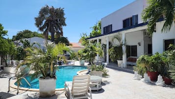 A pool area with lounge chairs, potted plants, and a white building with a tiled roof.