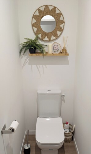 Een badkamer met een toilet, een spiegel aan de muur, een plankje met een potplant, een kaars en een dromenvanger.