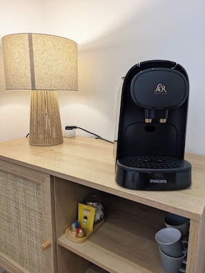 Une machine à café, une lampe et un petit meuble sur lequel se trouvent une tasse et un paquet, le tout posé sur une surface en bois.
