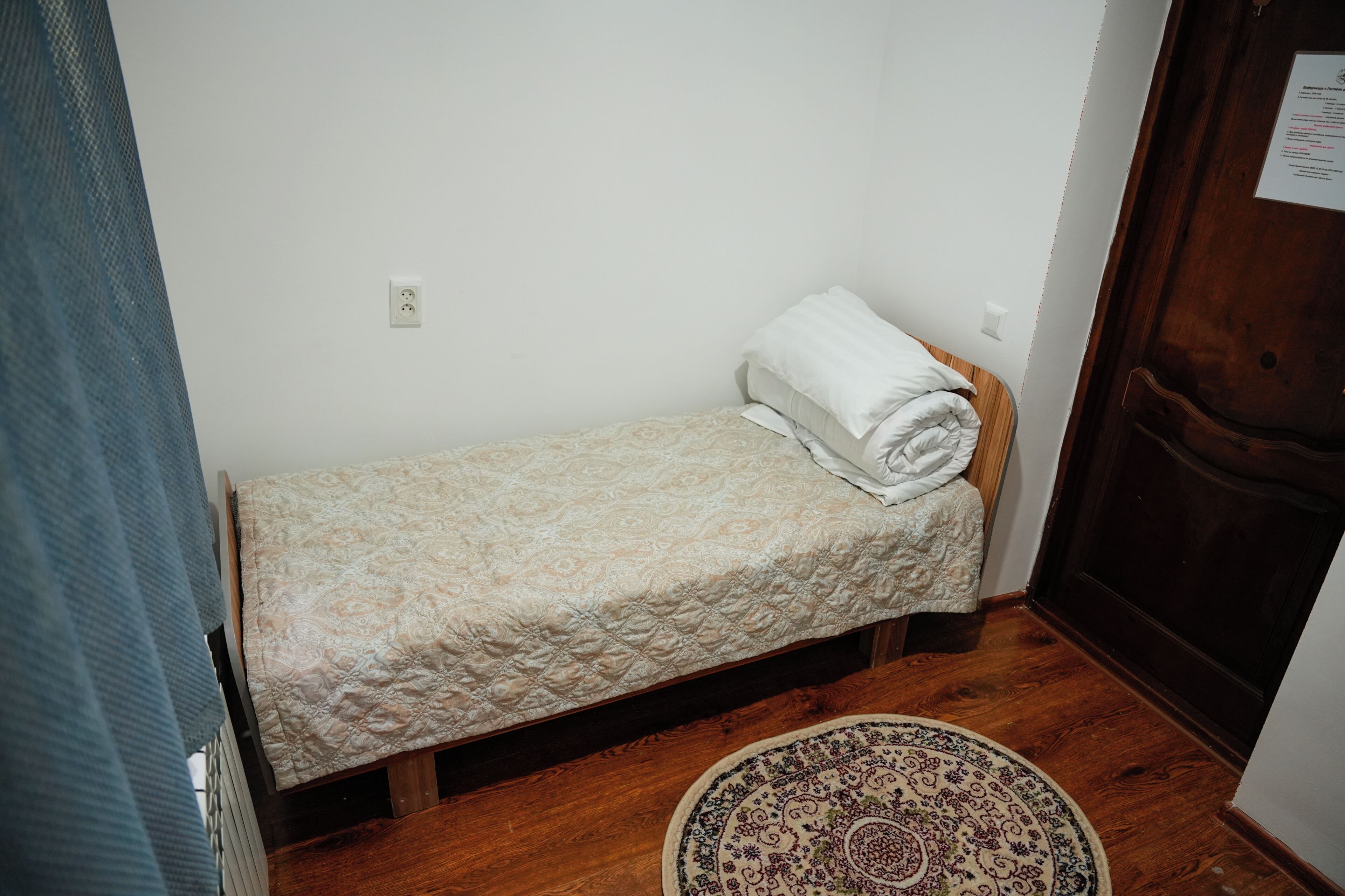 A small, single-bed room with a wooden floor, a patterned rug, and a door with a notice on it.