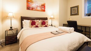 Una cama bien tendida con edredón blanco y almohadones decorativos, una mesita de noche con lámpara, y un cuadro en la pared.