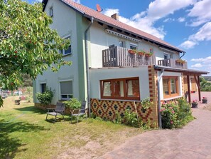 Ein zweistöckiges Gebäude mit Balkon, überdachter Terrasse und Gartenbereich mit Stühlen und Blumentöpfen.