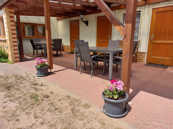 Ein überdachter Patio-Bereich mit Tisch, Stühlen, Topfpflanzen und einer Holztür.