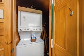 A compact laundry room with a washing machine and dryer, a hanging rack, and a basket with a box of laundry detergent. - Romantic Cabin * Hot tub * King Suite * Sauna * (Sevierville)