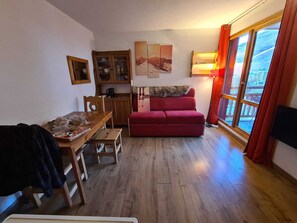 Ein gemütliches Wohnzimmer mit einem Holzesstisch, einem roten Sofa, einer Vitrine mit Glastüren und einem großen Fenster mit Vorhängen.