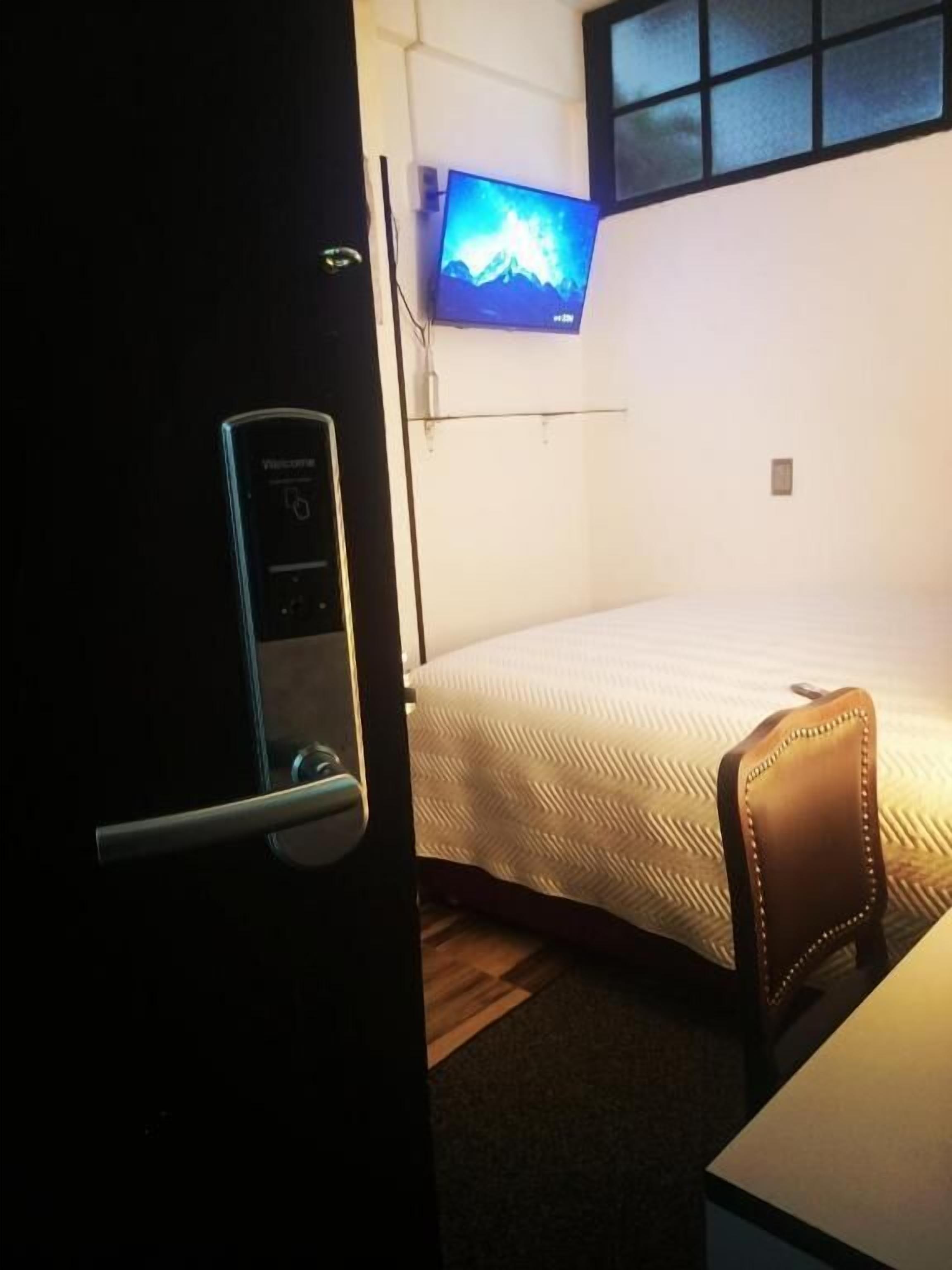 Habitación de hotel con cama, escritorio, silla, televisor montado en la pared y puerta con cerradura digital.