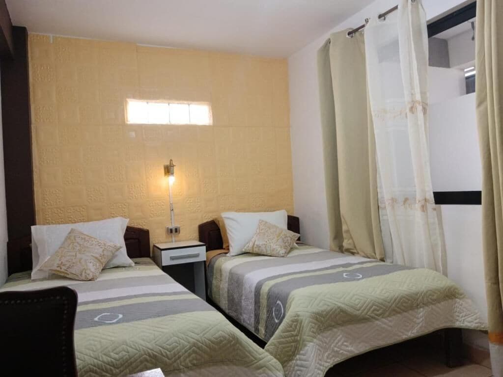 Une chambre d’hôtel avec deux lits, une table de chevet, une lampe fixée au mur et une fenêtre avec des rideaux.