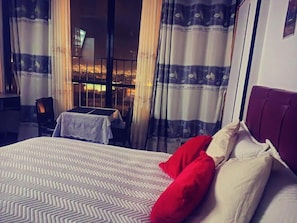A hotel room with a bed, a table, and a view of a cityscape at night.