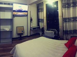 A hotel room with a bed, a wardrobe, a small table with a tablecloth, a picture frame, and a window with curtains.