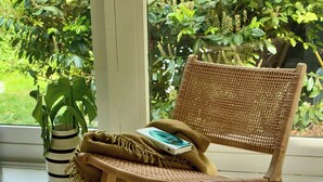 A wicker chair with a book and a blanket, a striped vase, and a window with greenery outside. - Authentic timber house with forest near Ebeltoft (Ebeltoft)