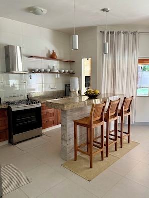A modern kitchen with a central island, bar stools, a stove, and a dining area with a view. - Arraial d'Ajuda House with Private Pool (Arraial d'Ajuda)
