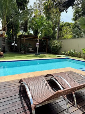 Piscina com espreguiçadeiras, rodeada por vegetação e um pérgula.