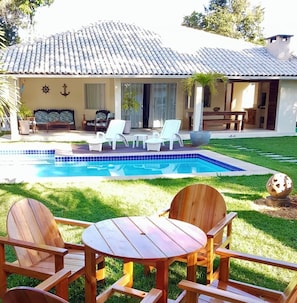 Uma área à beira da piscina com uma piscina azulejada, cadeiras de madeira e uma mesa.