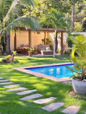 Piscina com borda em azulejo, área gramada com pedras no chão e área de estar coberta com almofadas e plantas.
