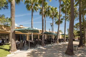 Un área de comedor al aire libre con mesas y sillas, rodeada de palmeras y cubierta con sombrillas.