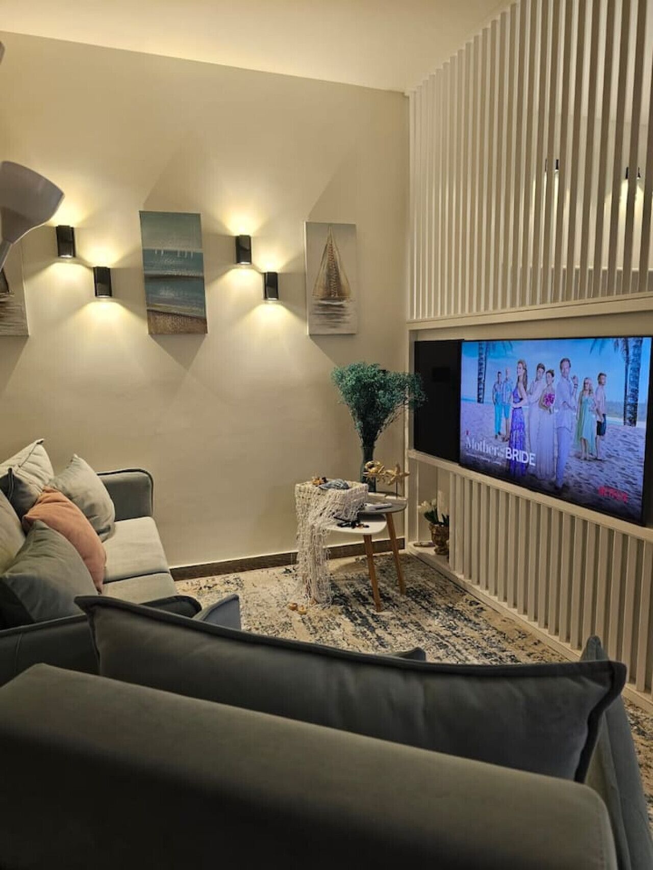 Ett modernt vardagsrum med en platt-TV, en grå soffa, väggmonterade vägglampor och en prydnadsväxt.