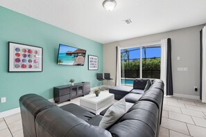 Ein modernes Wohnzimmer mit einem großen, grauen Ecksofa, einem weißen Couchtisch und einem an der Wand montierten Fernseher. Auf dem Boden befindet sich ein kleiner Schrank und ein Blumentopf.