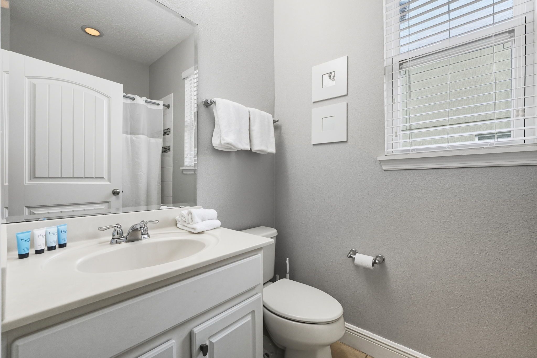Ein Badezimmer mit weißem Waschbecken, Spiegel und Toilette. Handtücher hängen an der Wand und ein Fenster mit Jalousien ist vorhanden.