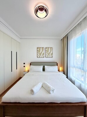 Ein Hotelzimmer mit Bett, Nachttischlampen, einem Fenster mit Vorhängen und zwei an der Wand befestigten Kunstwerken.