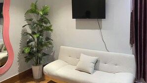 Uma sala de estar moderna com um sofá branco, uma televisão de tela plana na parede, um vaso de planta e piso de madeira.