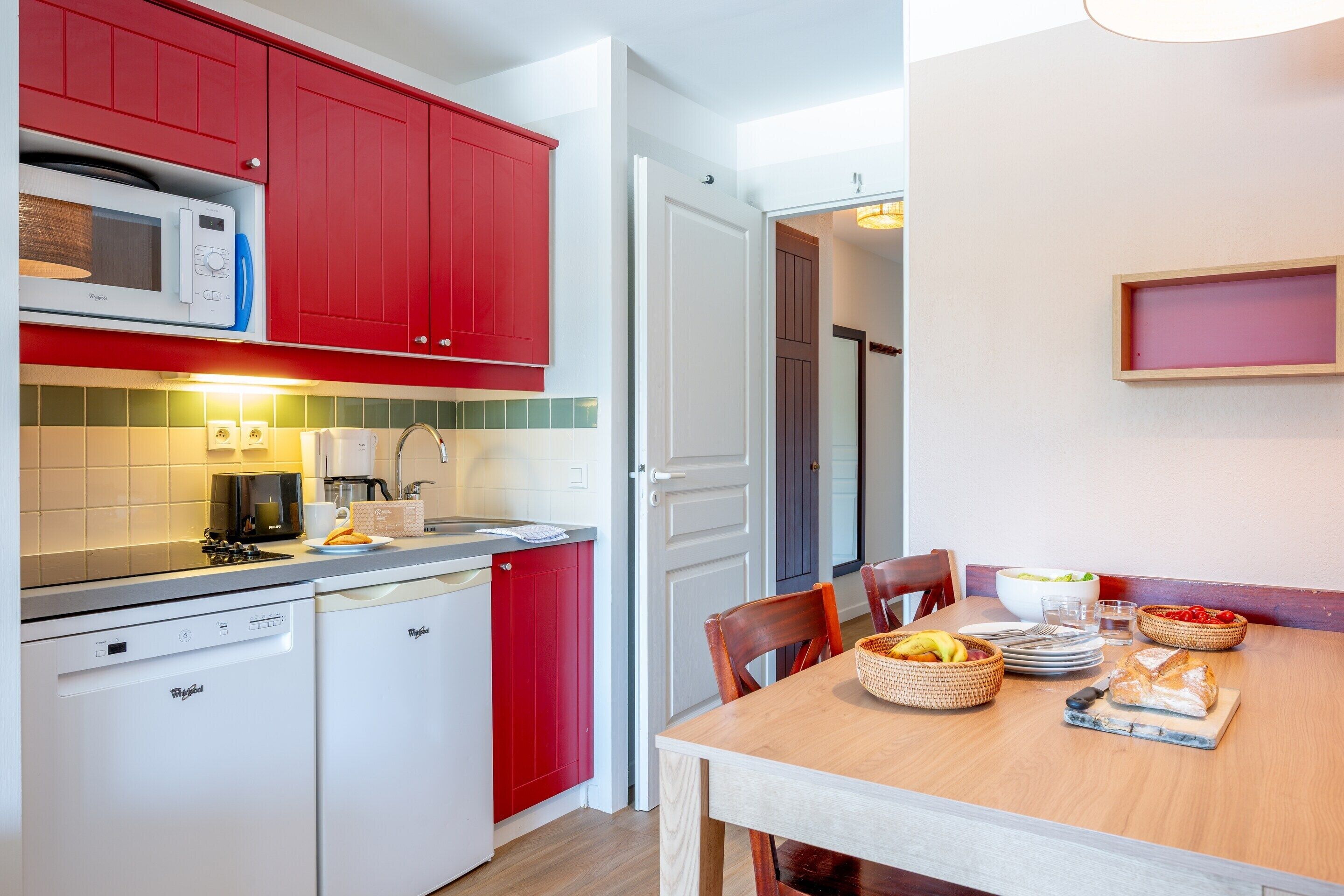 A kitchen with red cabinets, a microwave, a dishwasher, and a dining table set with plates, bowls, and a basket of fruit.