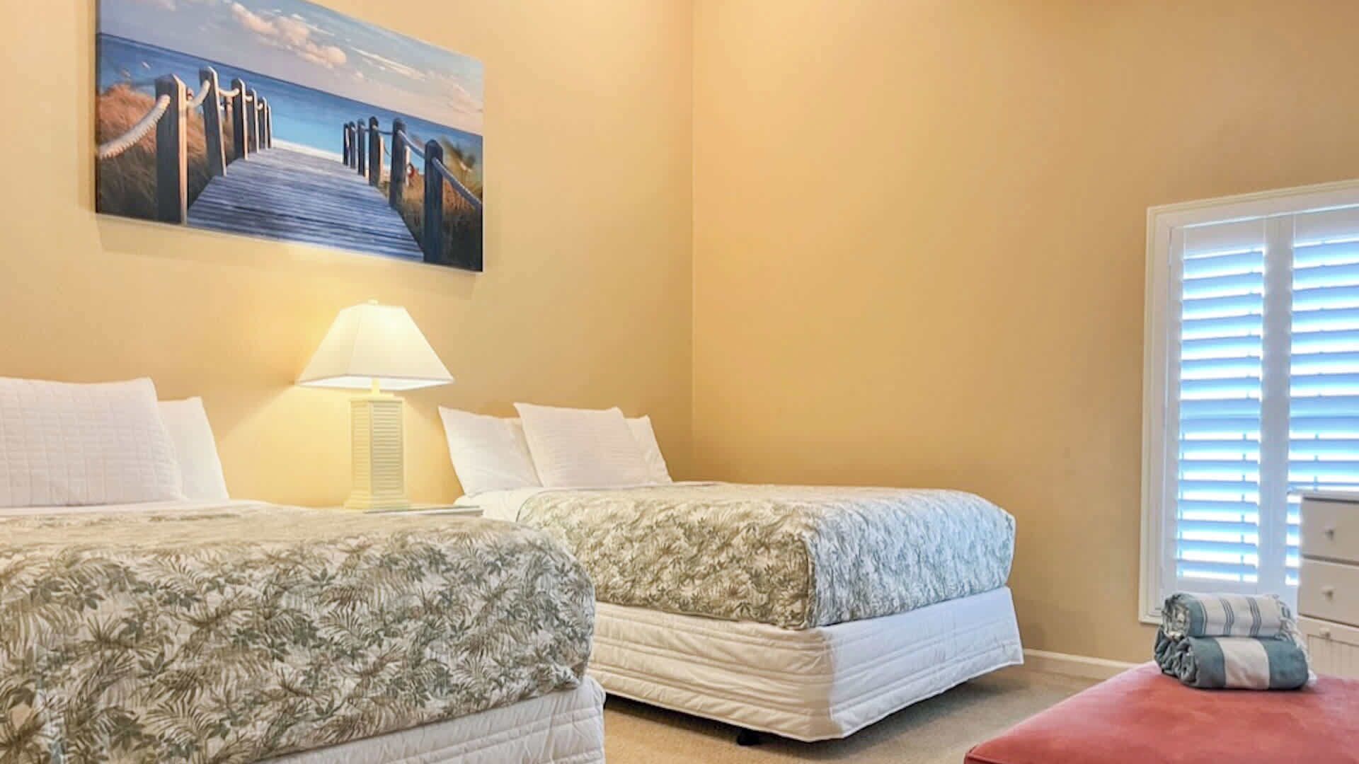 Une chambre avec deux lits, un tableau représentant une plage, une lampe et une fenêtre avec des stores.