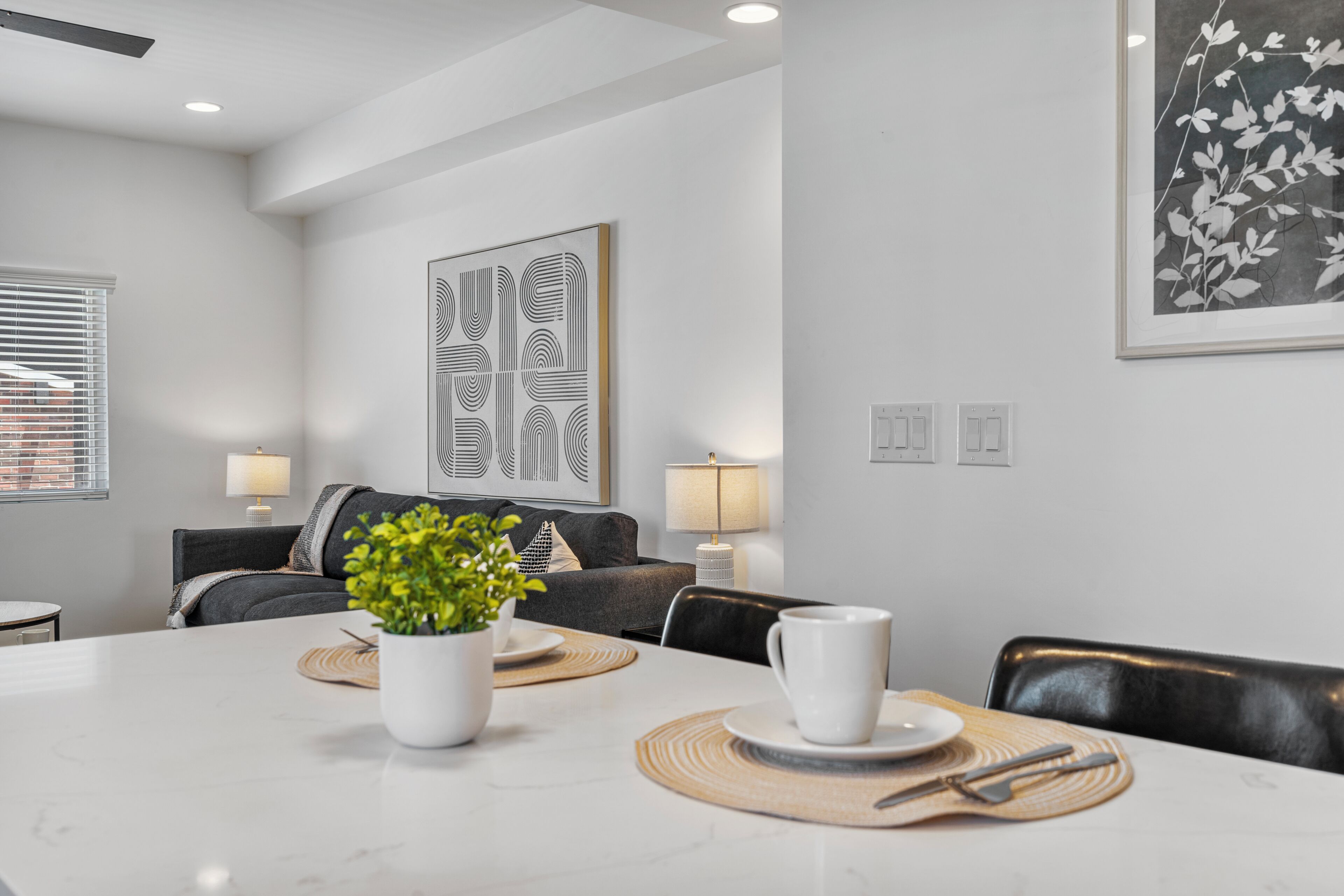A modern dining area with a white table set for two, a potted plant, and a large abstract artwork on the wall.