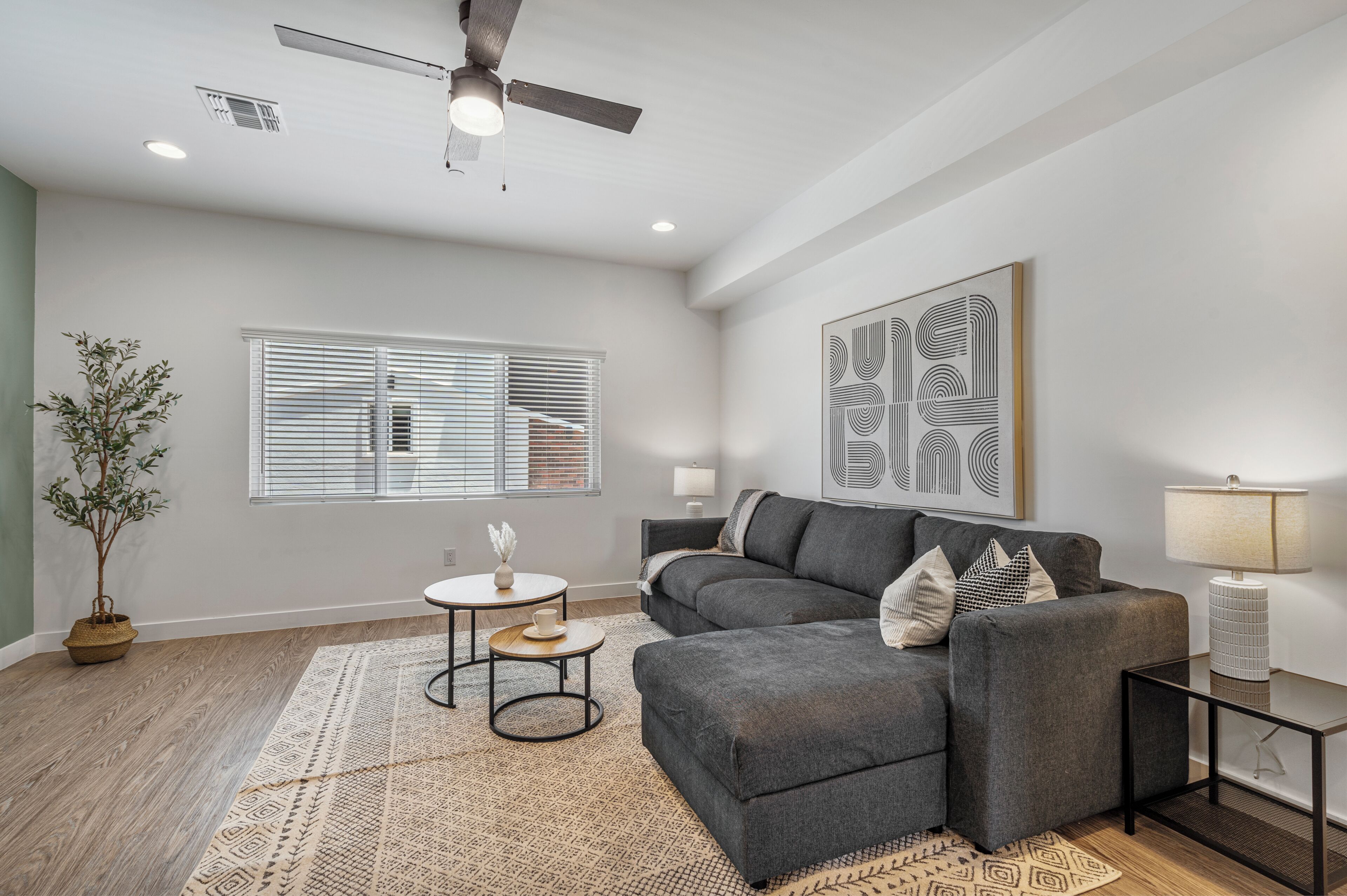 A modern living room with a grey sectional sofa, a small round table, a potted plant, and a ceiling fan.