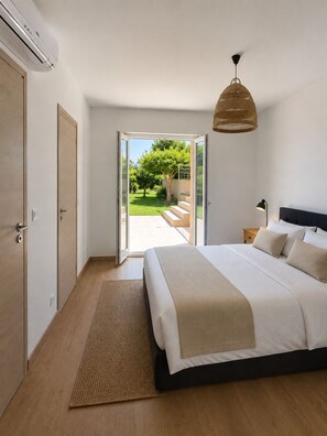 Ein Schlafzimmer mit Bett, Nachttisch, Lampe, einer Pendelleuchte und Blick auf einen Garten durch eine offene Tür.