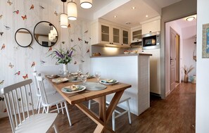 Un comedor con una mesa de madera, sillas blancas y un jarrón de flores. Se observan dos espejos redondos en la pared, y se distingue una cocina con armarios y un microondas al fondo.