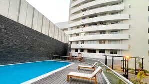 A swimming pool with wooden deck and lounge chairs, surrounded by a high-rise building with balconies. - Modern Apt in Lince Just Steps from San Isidro (Lince)