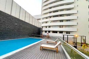 Ein Schwimmbecken mit Holzterrasse und Liegestühlen, umgeben von einem Hochhaus mit Balkonen.