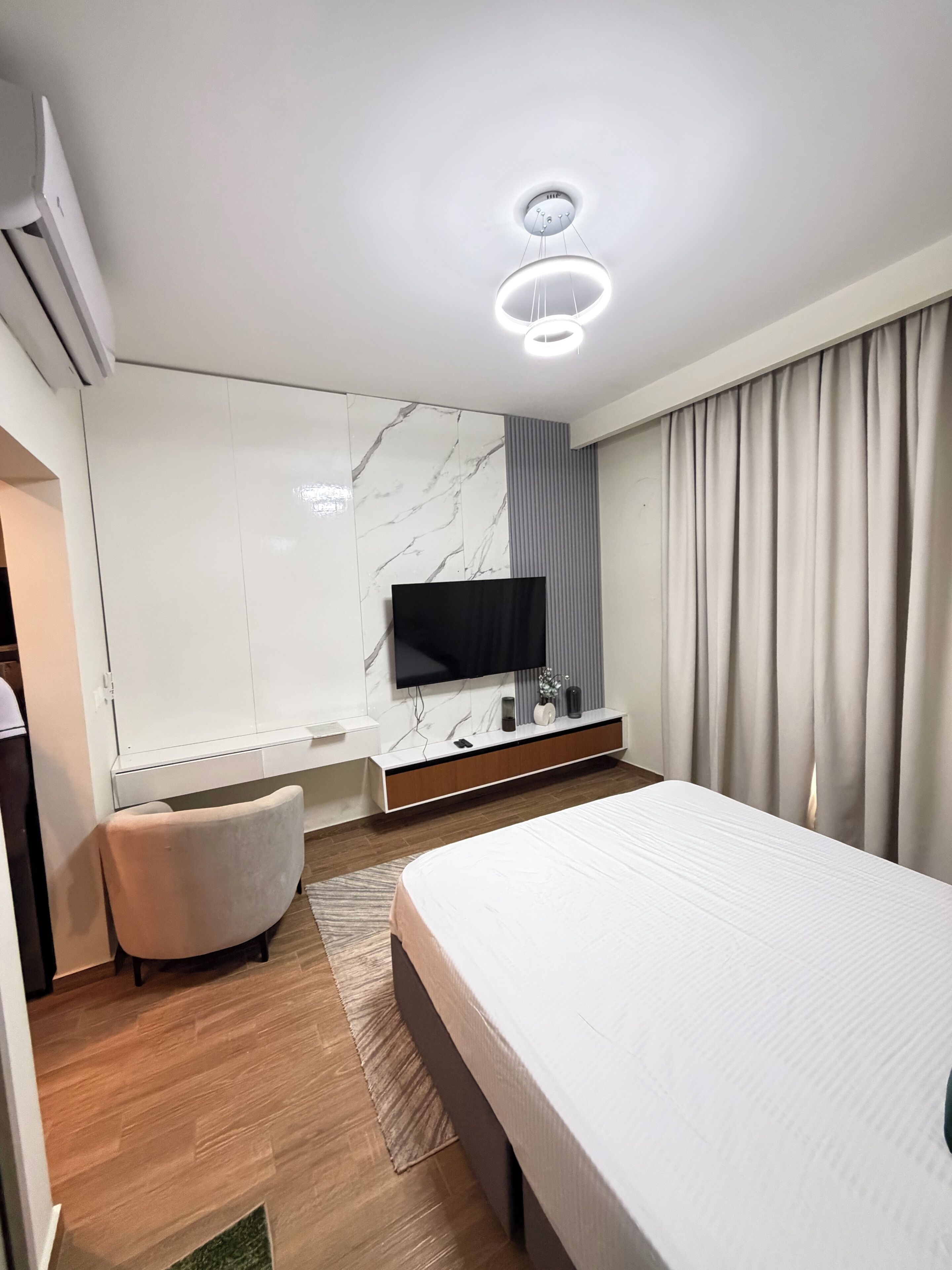 A modern hotel room with a large bed, a flat-screen TV mounted on a marble wall, a built-in air conditioning unit, and a large window with curtains.