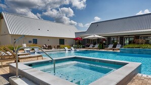 A swimming pool with lounge chairs and umbrellas, a covered area with a metal roof, and a building with a white facade and a blue sign. - Landing | Spectacular 1BD, Gym, Pool (Madison)