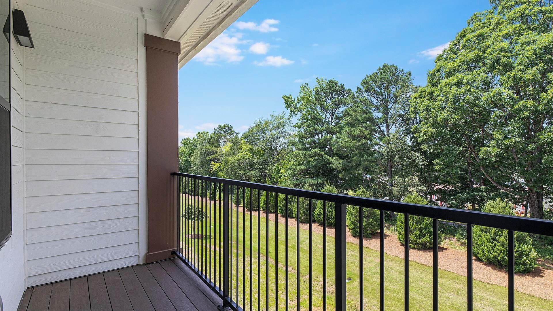 A balcony with a wooden floor and a black railing, overlooking a lawn with trees and a clear sky.