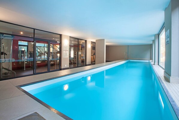 Une piscine intérieure avec des murs en verre, une eau d’un bleu limpide et, au fond, on peut apercevoir des appareils de musculation.
