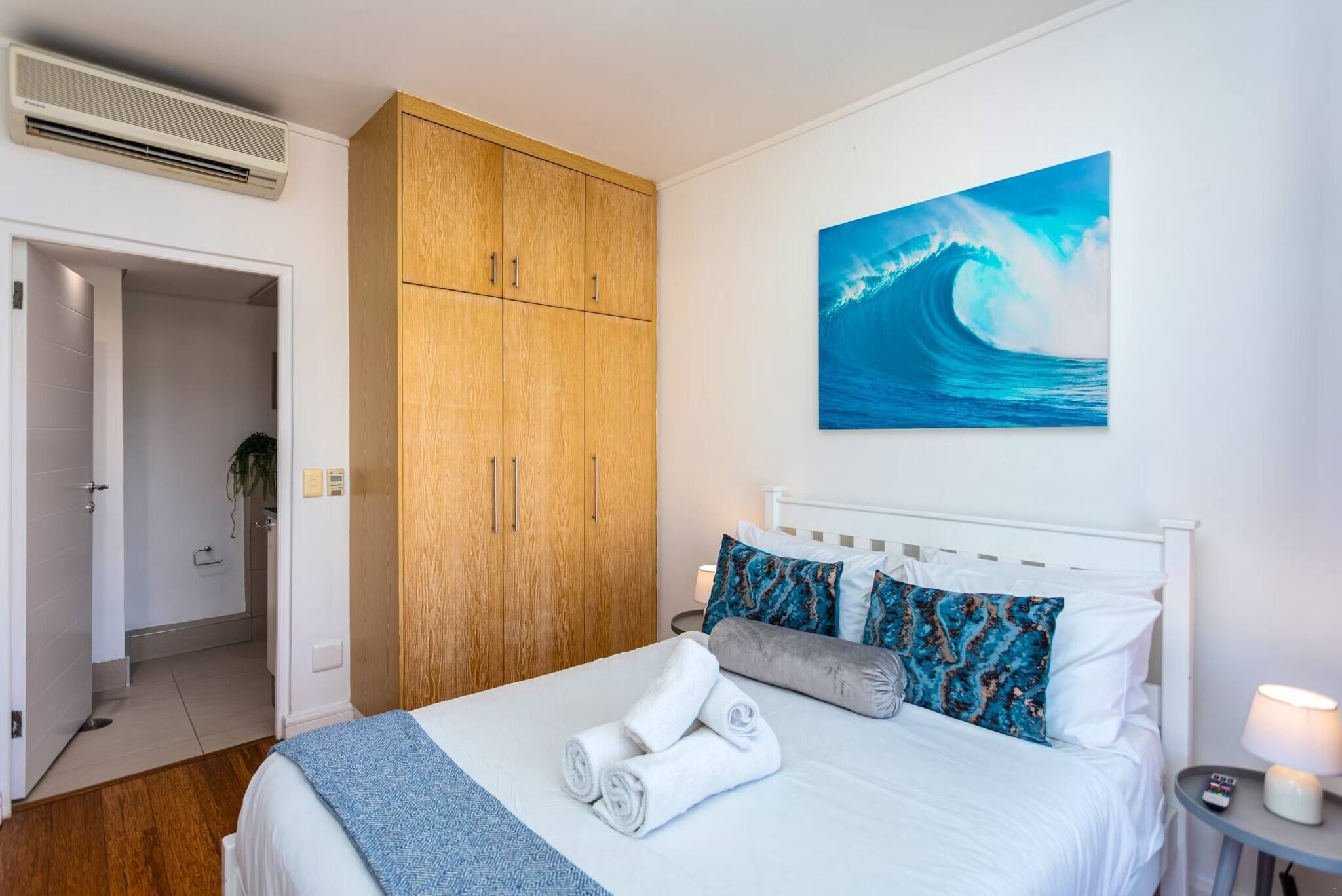Ein Hotelzimmer mit einem Bett, einem Holzschrank, einem Gemälde einer Welle und einem durch eine offene Tür einsehbaren Badezimmer.