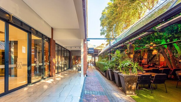 A covered walkway with glass storefronts, outdoor seating, and greenery. - Mountain Views 2BR Furnished De Waterkant (Cape Town)