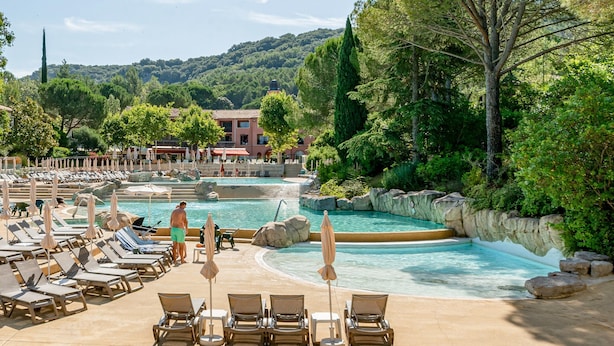 Zona de piscina con sillas de descanso y sombrillas, rodeada de follaje y edificios.