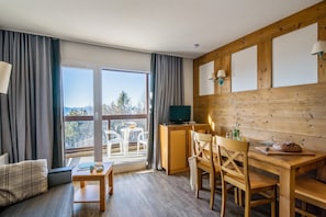 A room with a wooden desk, a television, a dining table with chairs, and a balcony with a view. - Pyrenees Alpine Apartment for 5 (Font-Romeu-Odeillo-Via)