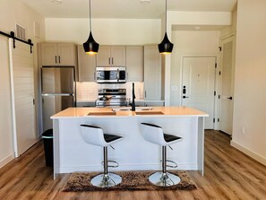 A modern kitchen with a central island, two bar stools, a stainless steel refrigerator, a microwave, and an oven.
