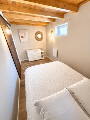 A bedroom with a wooden ceiling, a bed with floral bedding, a dresser, a wall-mounted mirror, and a window.