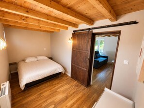 A bedroom with a bed, wooden flooring, a ceiling with exposed beams, a sliding door, and a view to a balcony.
