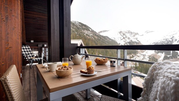 A wooden table with breakfast items, a wicker chair, and a view of snow-capped mountains. - Mountainside Suite With Hot Tub (Araches-la-Frasse)