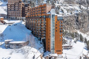 Un hotel de varios pisos con balcones, una pista de esquí y un paisaje montañoso de fondo.