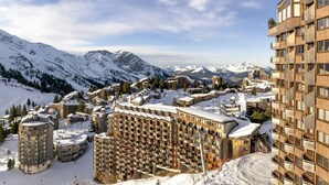Eine verschneite Bergstadt mit mehrstöckigen Gebäuden, darunter ein Hotel mit Balkonen, und einem Skiort im Hintergrund.