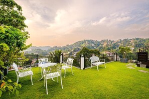 Un patio con sillas y mesas de metal blanco, un césped verde y vistas a un paisaje montañoso.