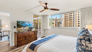 Habitación de hotel con cama, ventilador de techo, televisor, cómoda y vistas a edificios a través de la ventana.