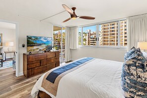 Ein Hotelzimmer mit einem Bett, einem Deckenventilator, einem Fernseher, einem Schminktisch und Blick auf Gebäude durch das Fenster.