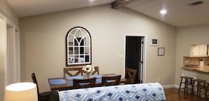 Comedor con mesa, sillas, un florero con flores, una ventana y una zona de cocina con taburetes.